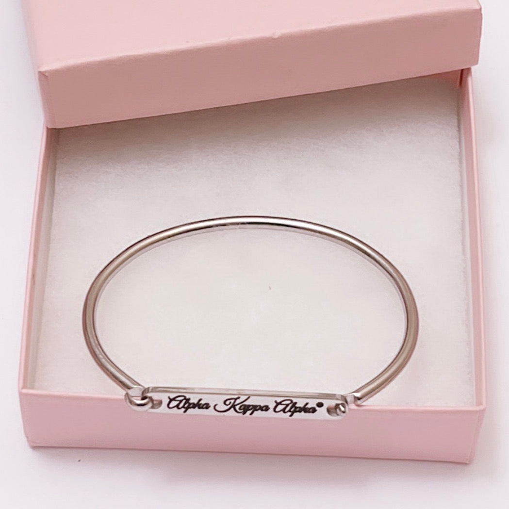 A polished stainless steel bracelet bar cuff engraved with the words “Alpha Kappa Alpha®.” The elegant bracelet rests inside a light pink gift box with a soft white cushioned interior, highlighting its sleek and minimalist design.