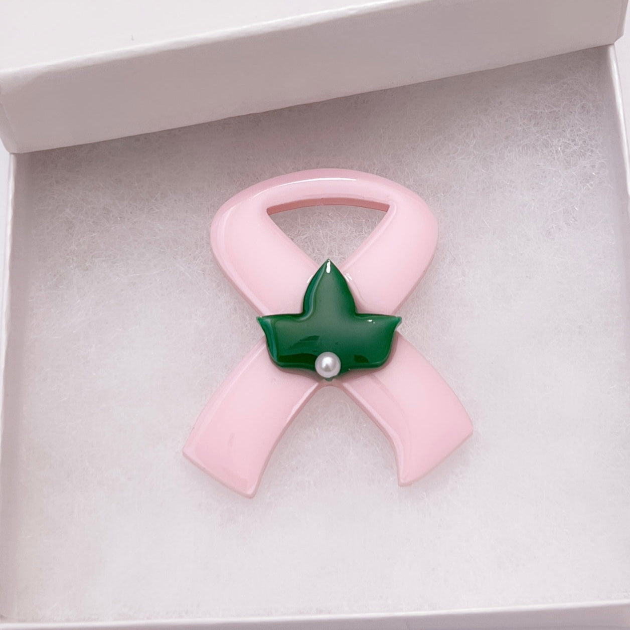 Pink ribbon brooch with green leaf and pearl center on a white gift box.