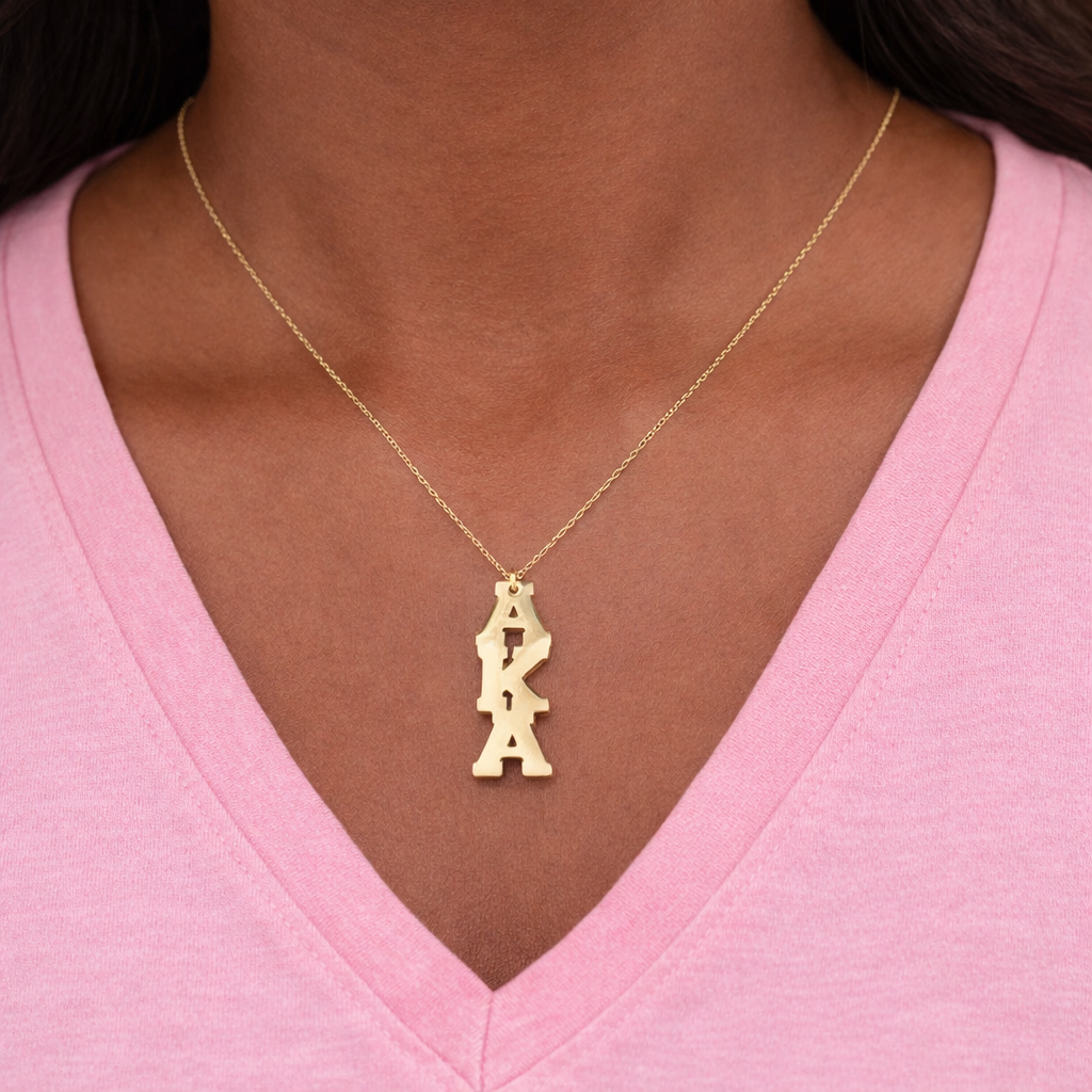 Gold necklace with 'AKA' pendant on a person wearing a pink shirt