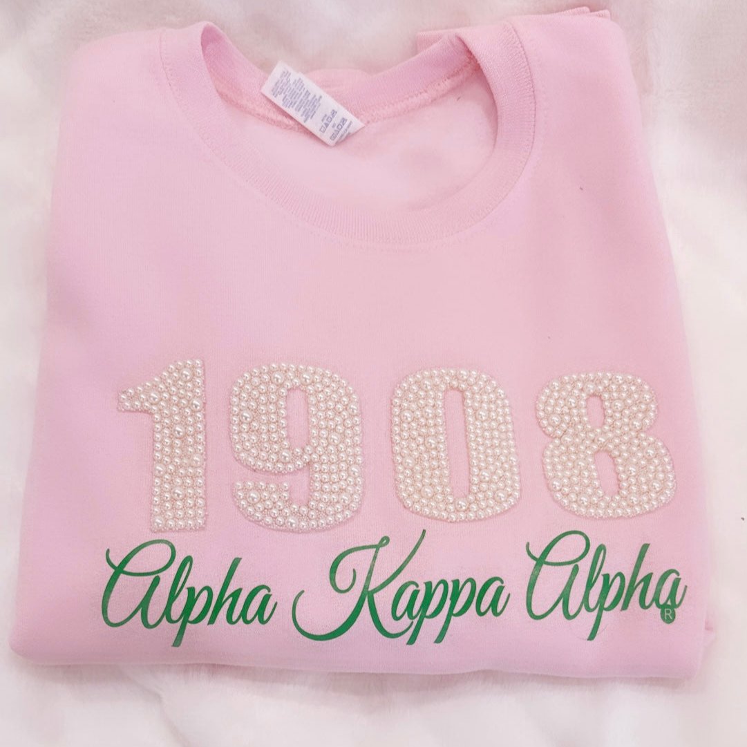 "Pink sweatshirt adorned with '1908' in pearls and 'Alpha Kappa Alpha' in green lettering, perfect for members of Alpha Kappa Alpha Sorority, Inc., to showcase their pride in comfort and style."
