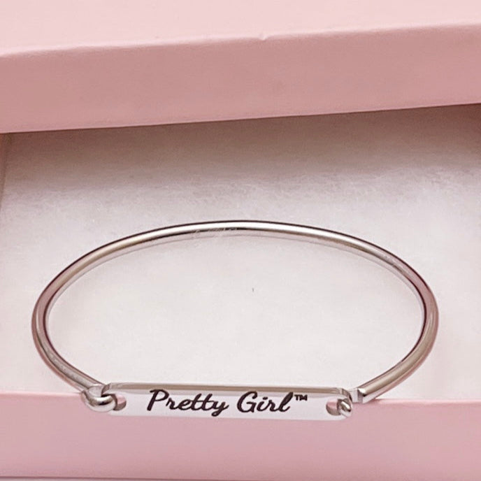 A stainless steel bracelet bar cuff engraved with the words “Pretty Girl™.” The elegant bracelet is displayed inside a light pink gift box with a white cushioned interior, emphasizing its simple and polished design.