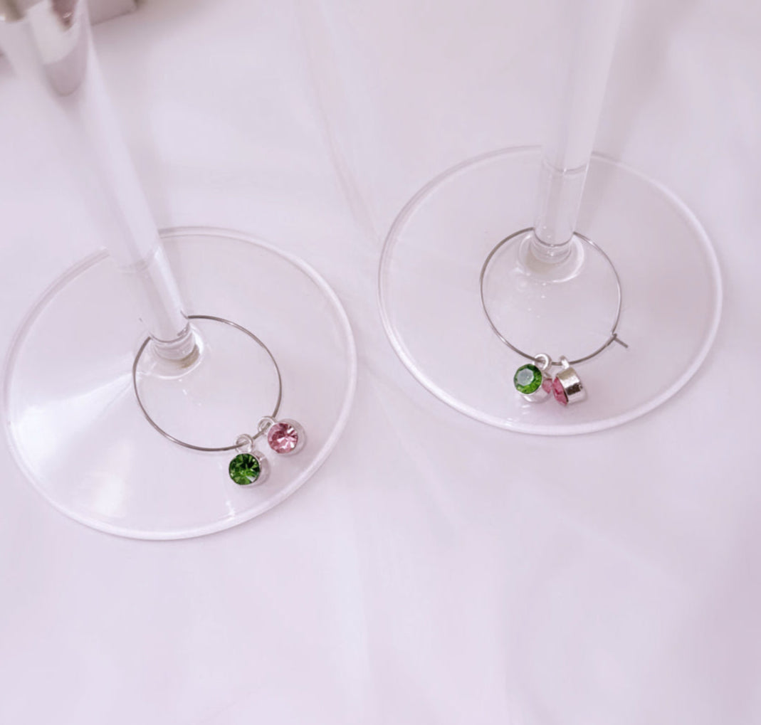 Close-up of wine glass stems with silver-tone charms featuring pink and green crystals, used as wine glass markers for sorority gatherings and celebrations.