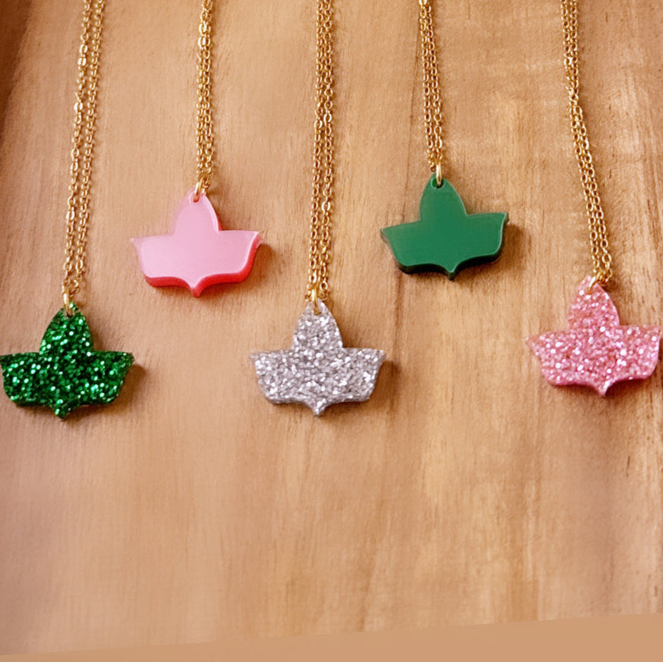 Five ivy leaf pendant necklaces with gold chains displayed on a wooden surface. The pendants include green glitter, solid pink, silver glitter, solid green, and pink glitter finishes.