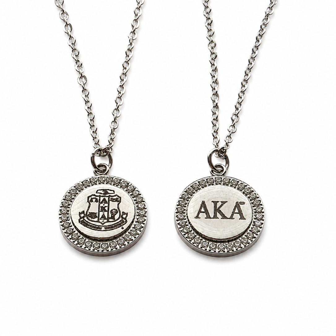 Two silver chain necklaces with round pendant charms on a white background. One pendant features a crest design, and the other is engraved with the letters 'AKA' inside a textured bezel.