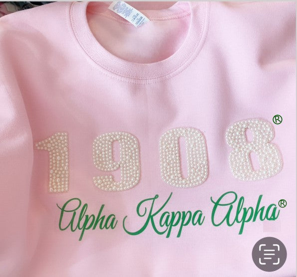 Pink folded sweatshirt in pink and green, featuring "1908" embellished with pearls and "Alpha Kappa Alpha" written in elegant script, representing Alpha Kappa Alpha Sorority, Inc.®️.