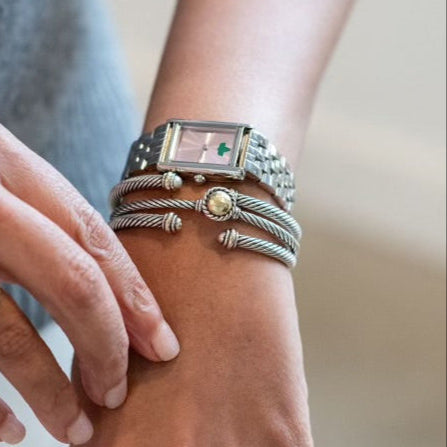 Close-up of a woman’s wrist adorned with a rectangular silver watch from The Timeless Collection. The watch features a soft pink dial with a green ivy emblem, paired with elegant silver bangles for a polished and sophisticated look.