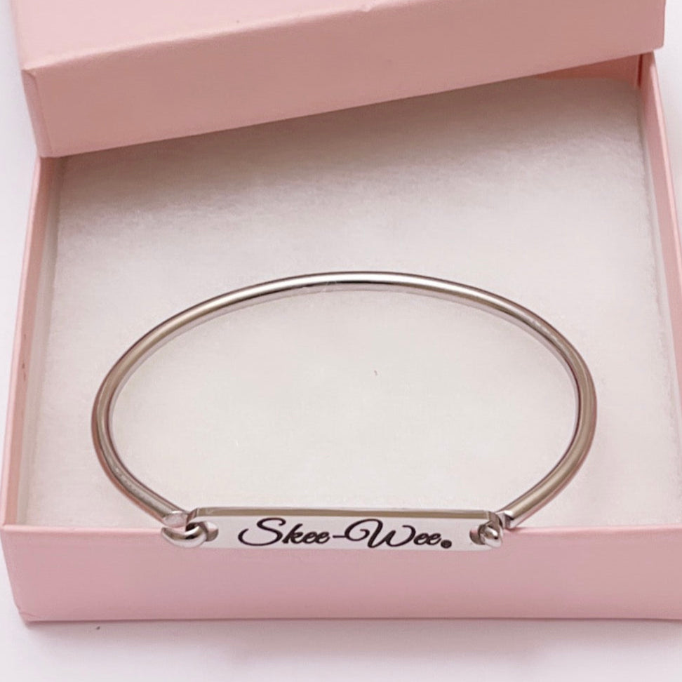 A sleek stainless steel bracelet bar cuff engraved with the words "Skee-Wee." The bracelet is nickel-free, hypoallergenic, and presented in a soft pink gift box with a white cushioned insert, showcasing it's elegant and polished design. 