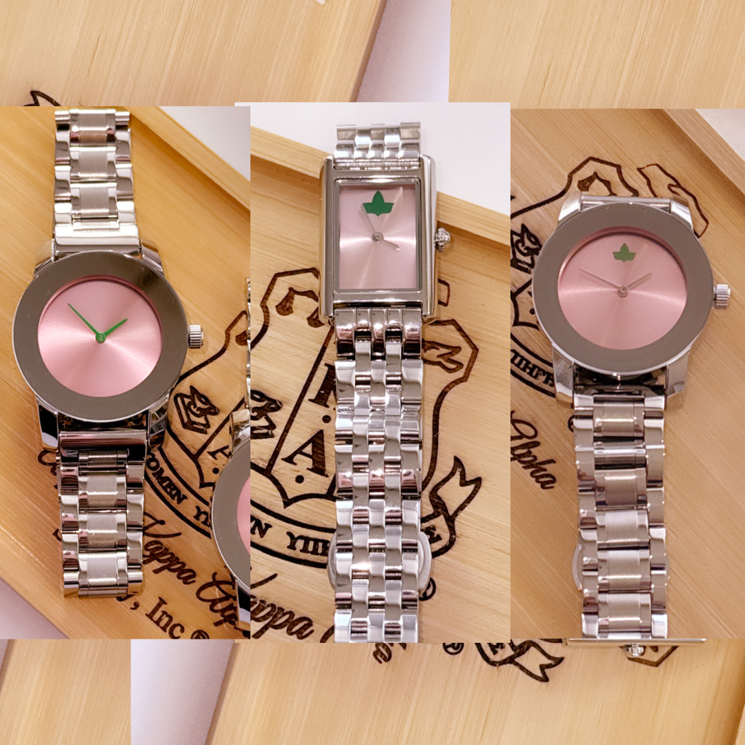 Three elegant watches from The Timeless Collection displayed on a wooden surface engraved with the Alpha Kappa Alpha Sorority, Inc. crest. Each watch features a stainless steel band, soft pink sunburst dial, and green ivy accents, showcasing timeless style and sisterhood-inspired design.