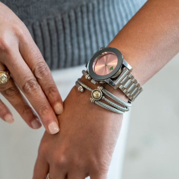 Close-up of a woman’s wrist wearing a stylish silver watch from The Timeless Collection. The watch features a soft pink sunburst dial with a green ivy emblem, paired with elegant silver bangles, showcasing a chic and sophisticated accessory combination.