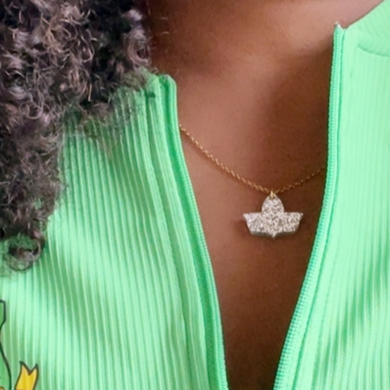 Close-up of a woman wearing a sparkly ivy leaf pendant on a gold chain, paired with a ribbed green top featuring the Alpha Kappa Alpha Sorority, Inc.® crest.