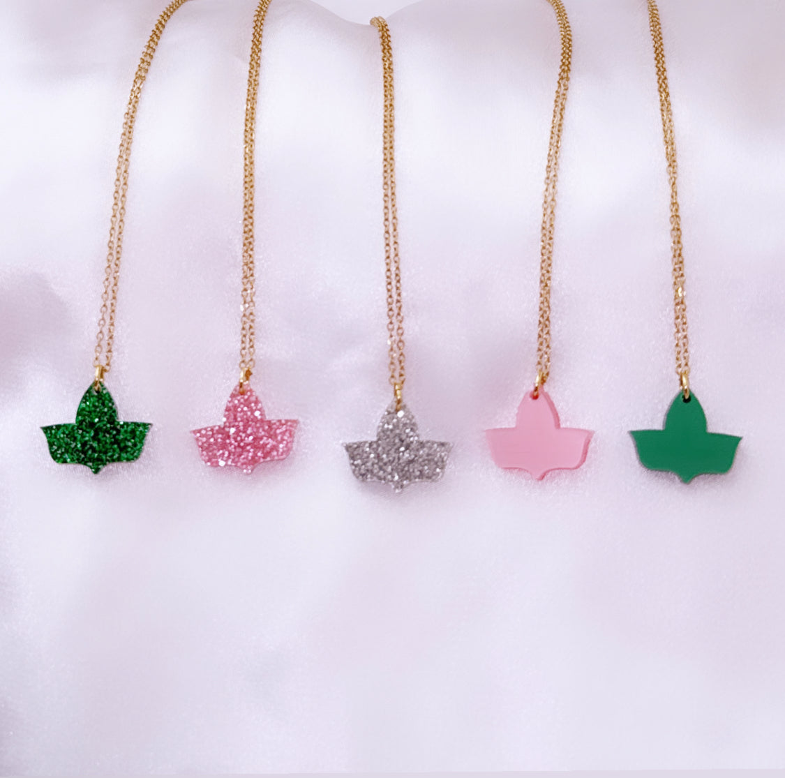 Five ivy leaf-shaped pendant necklaces on gold chains are displayed on a white satin surface. The pendants feature different finishes: green glitter, pink glitter, silver glitter, solid pink, and solid green.