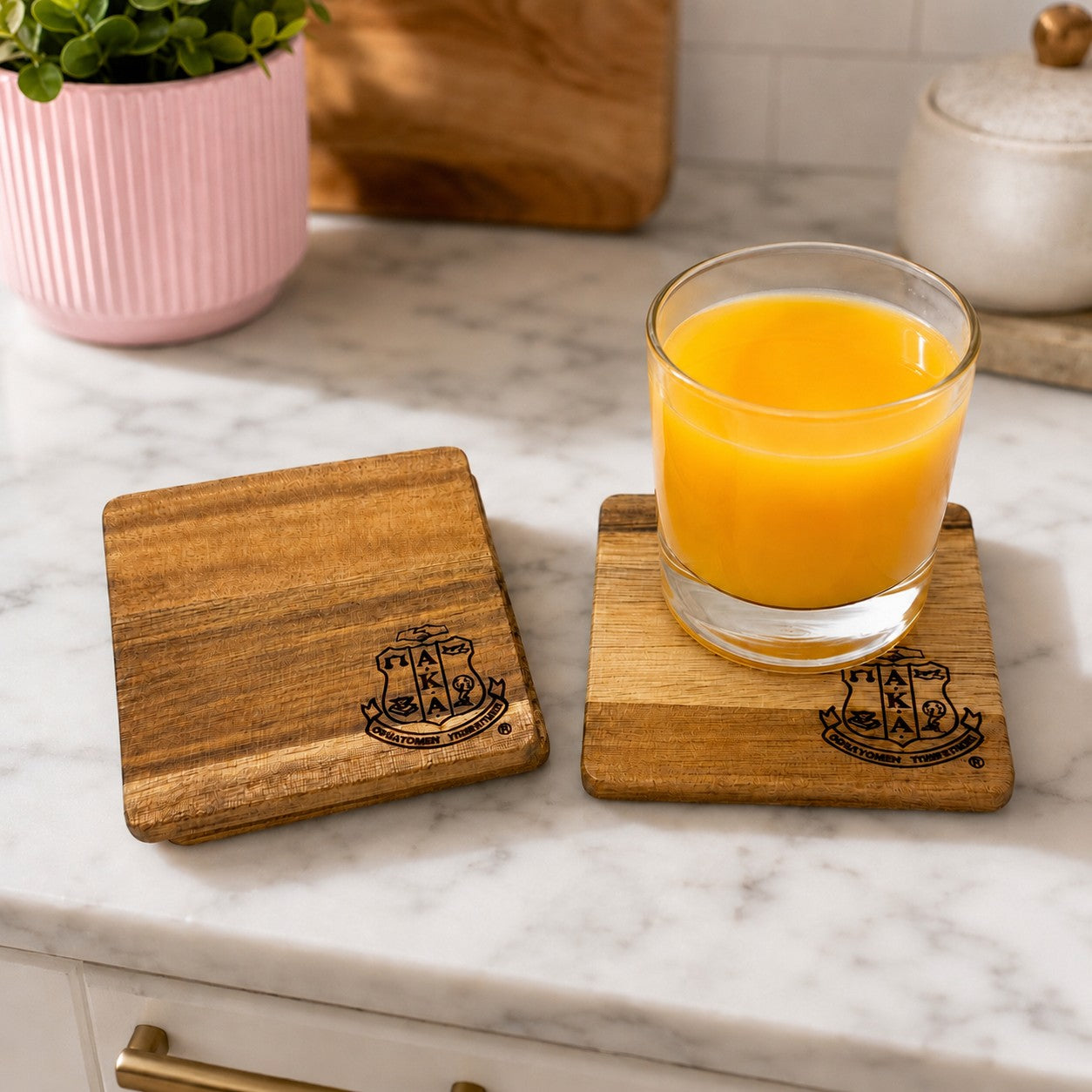 Lifestyle image of Alpha Kappa Alpha Sorority, Inc.® engraved acacia wood coasters displayed on a marble kitchen countertop with a glass of orange juice and a soft pink planter, showcasing the natural wood grain and elegant everyday home décor