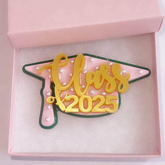 Multi-layer acrylic graduation cap brooch with pink and green base, pearl embellishments, and gold "Class of 2025" text, displayed in a pink gift box. 