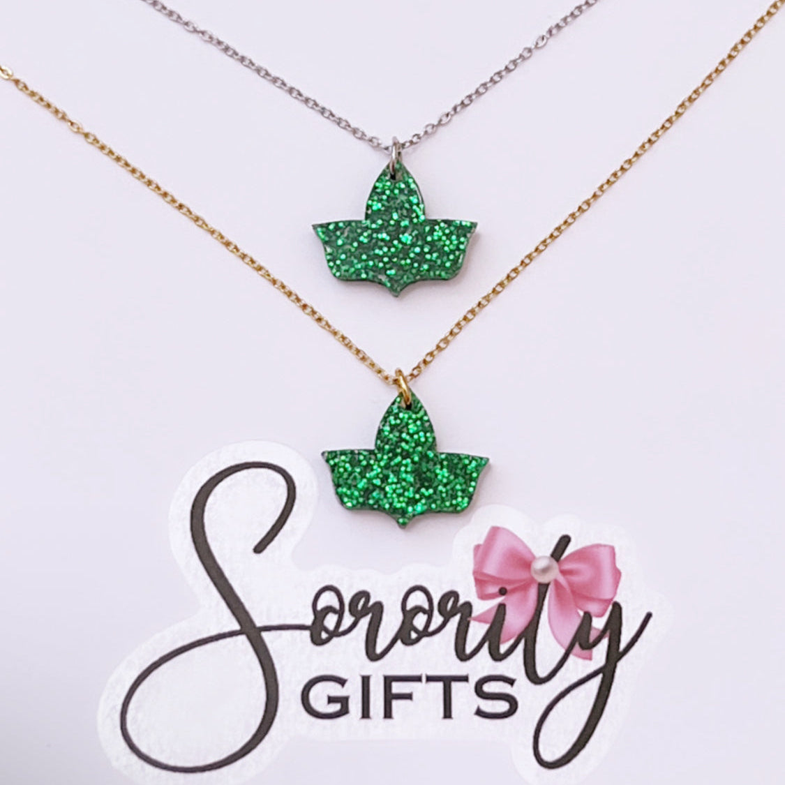Two green glitter ivy leaf pendant necklaces displayed on a white card with the "Sorority Gifts" logo. One necklace features a silver chain, and the other has a gold chain.
