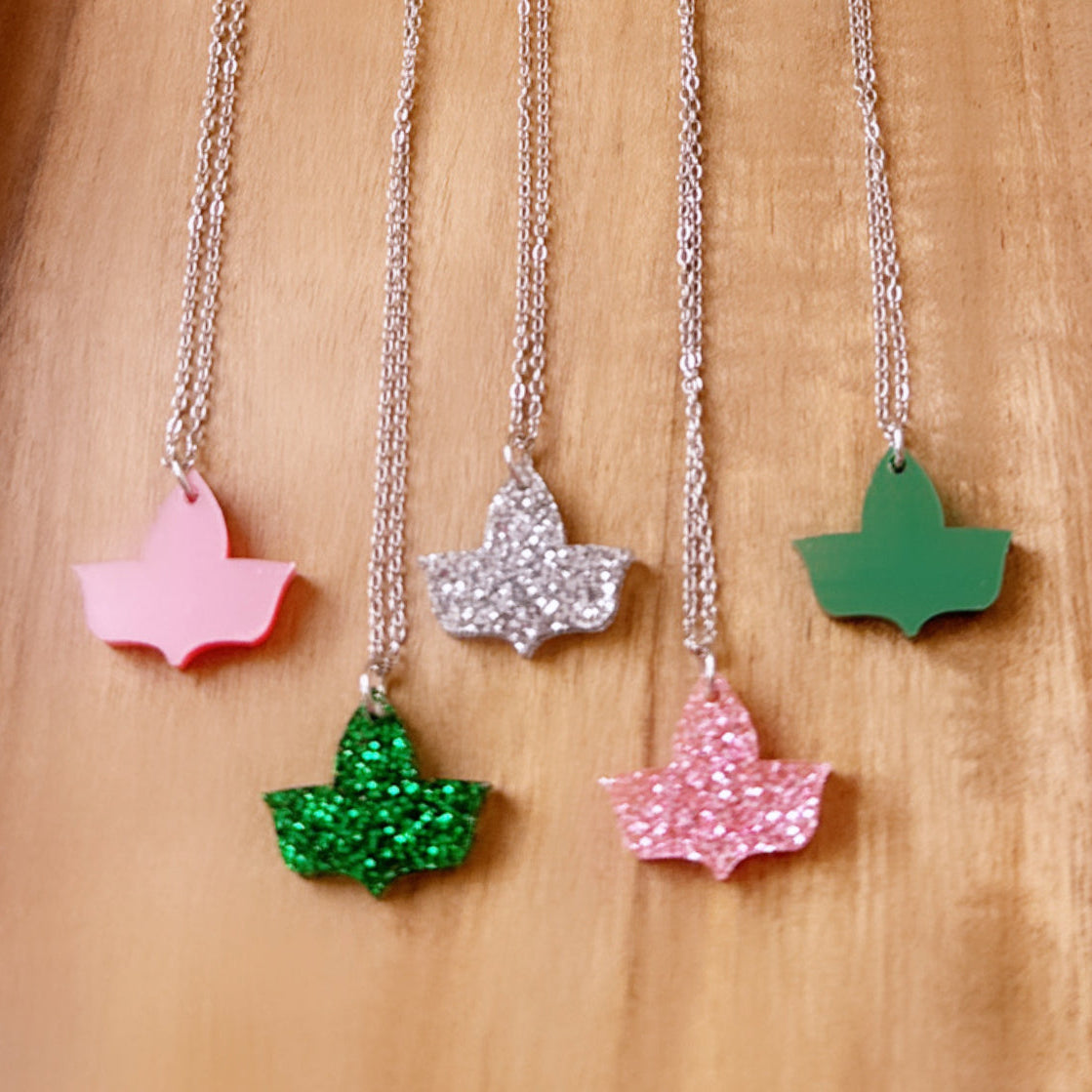 Five ivy leaf pendant necklaces with silver chains displayed on a wooden surface. The pendants include finishes in solid pink, green glitter, silver glitter, pink glitter, and solid green.