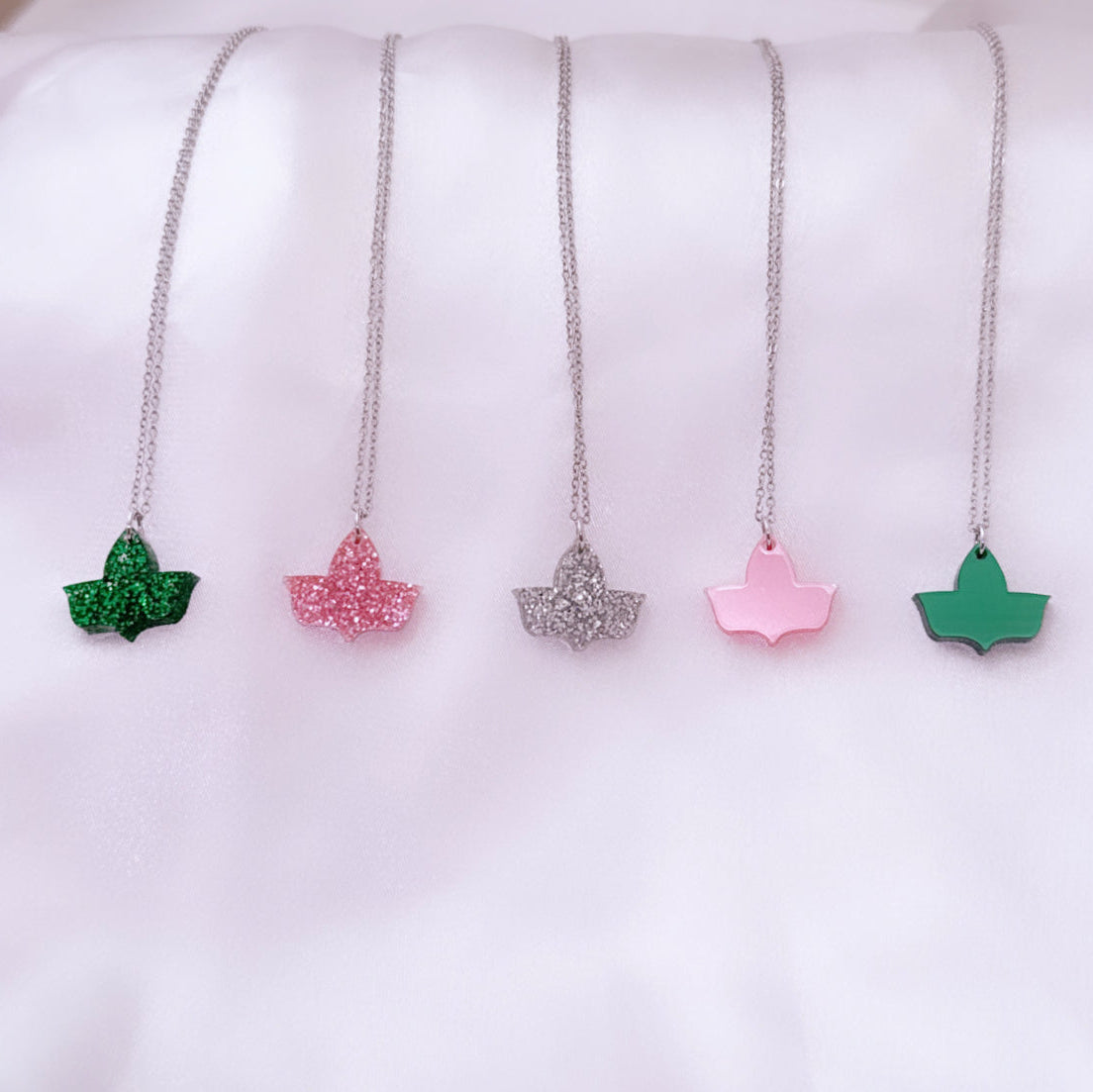 Five ivy leaf pendant necklaces with silver chains displayed on a soft white satin surface. The pendants feature different finishes: green glitter, pink glitter, silver glitter, solid pink, and solid green.