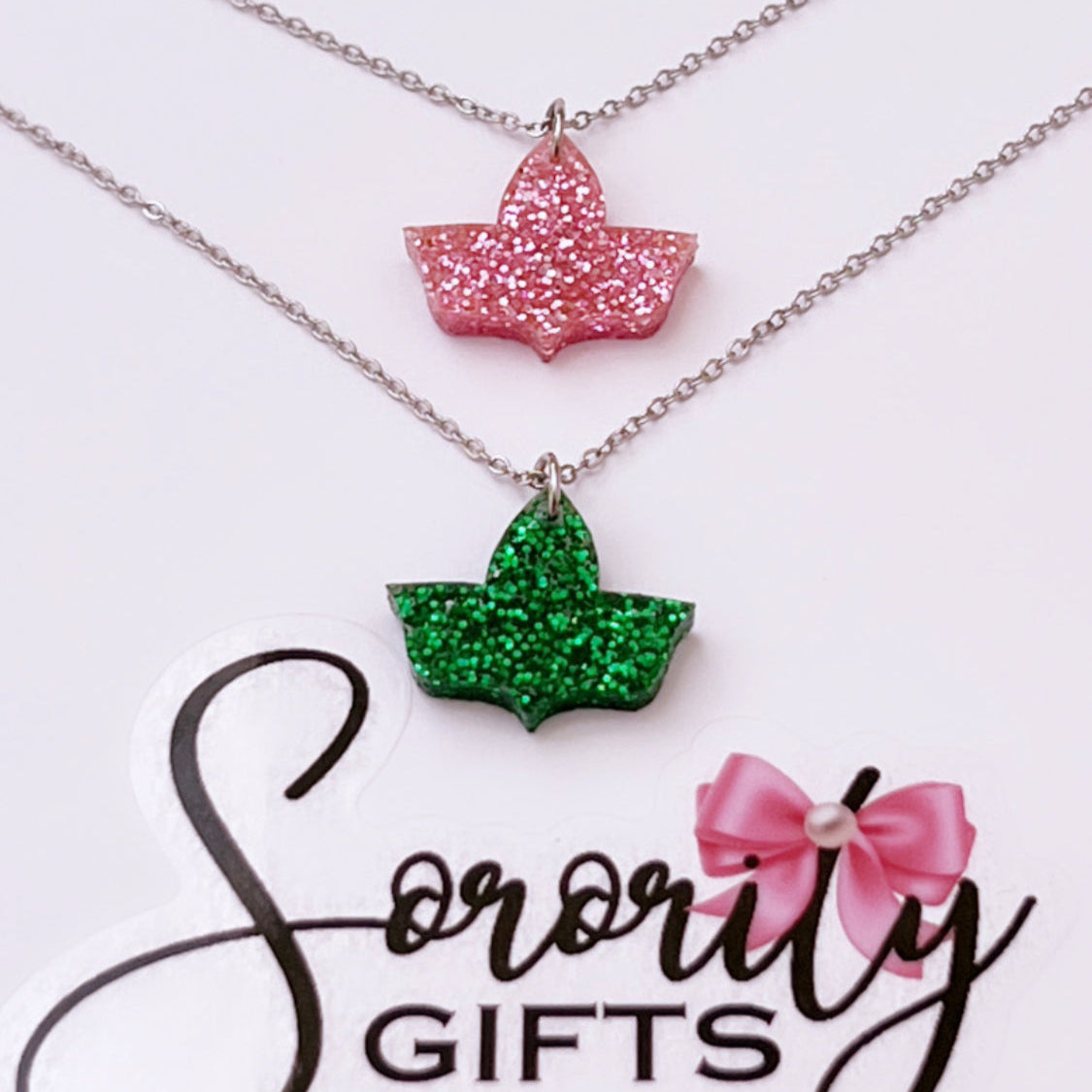 Two ivy leaf pendant necklaces with silver chains are displayed—one in pink glitter and the other in green glitter—on a white card featuring the "Sorority Gifts" logo with a pink bow.