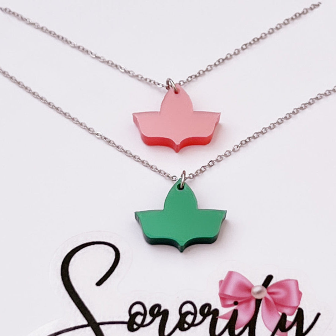 Two ivy leaf pendant necklaces with silver chains are displayed—one in solid pink and the other in solid green—on a white card with the "Sorority Gifts" logo featuring a pink bow.